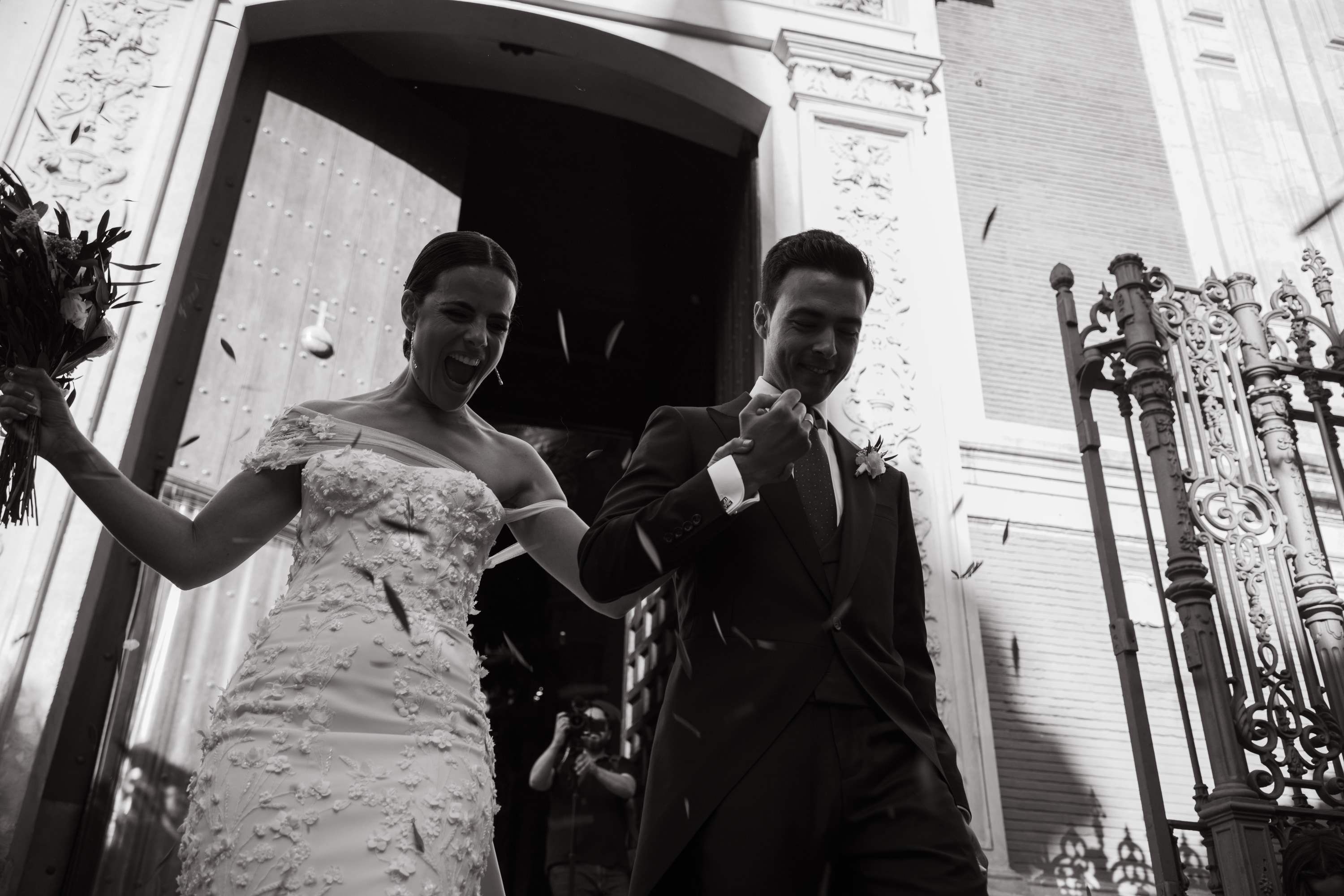 Los novios salen de la iglesia de la mano, riendo, bajo una lluvia de pétalos